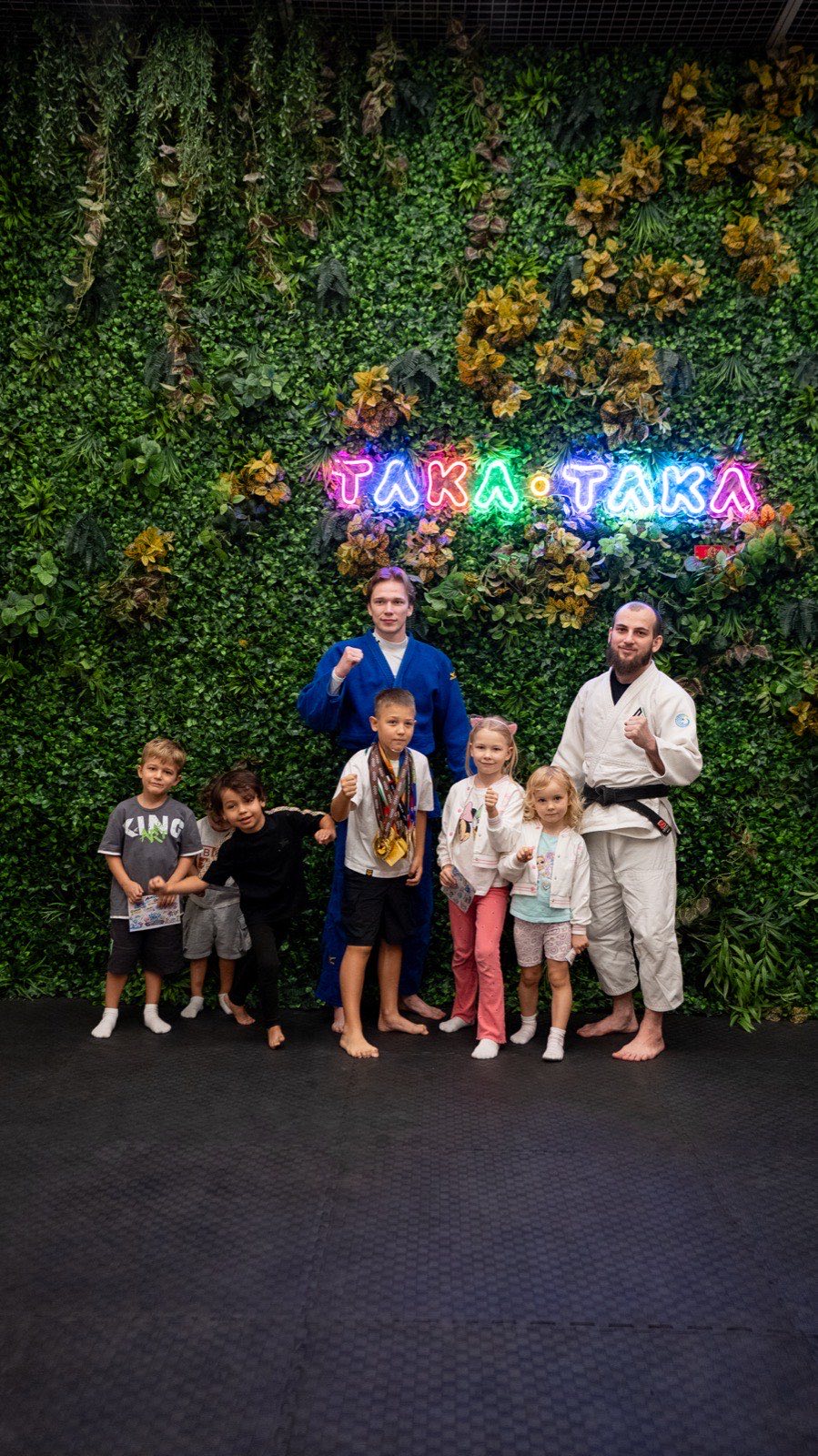 Children with two jiu-jitsu coaches at the green wall under the club sign